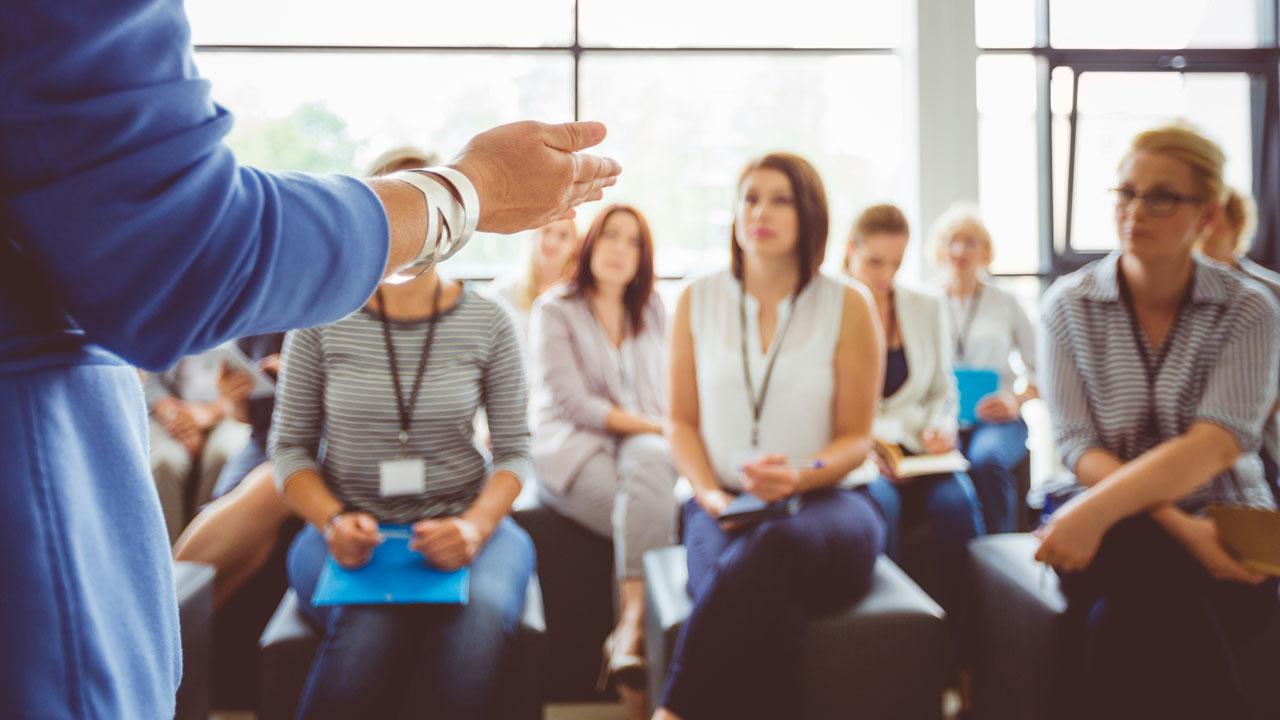 A blurred view of people sitting in a casual classroom, facing a presenter wearing a blue jacket whose arm and side are shown in the foreground